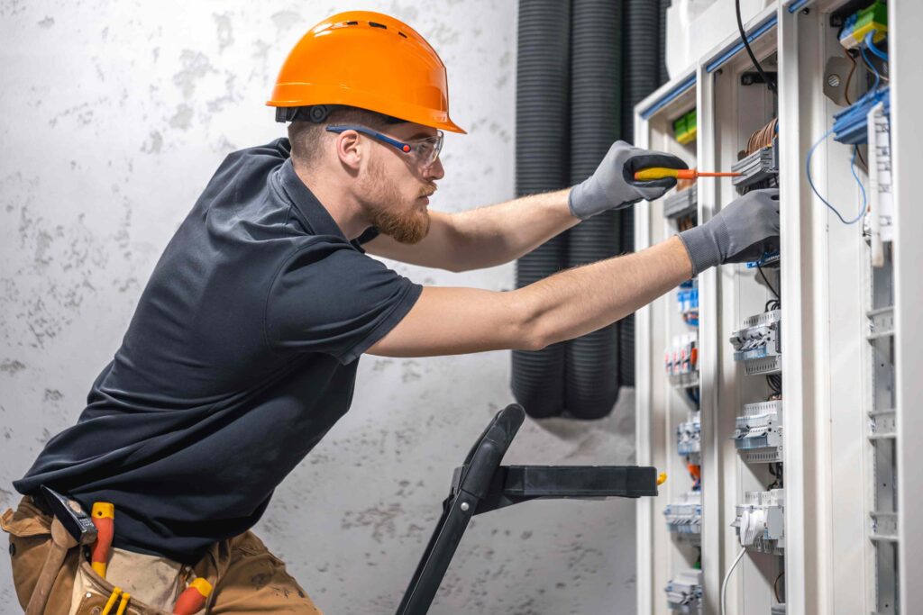 Elektriker mit Schutzhelm arbeitet an einem Schaltschrank mit Kabeln und Werkzeug.
