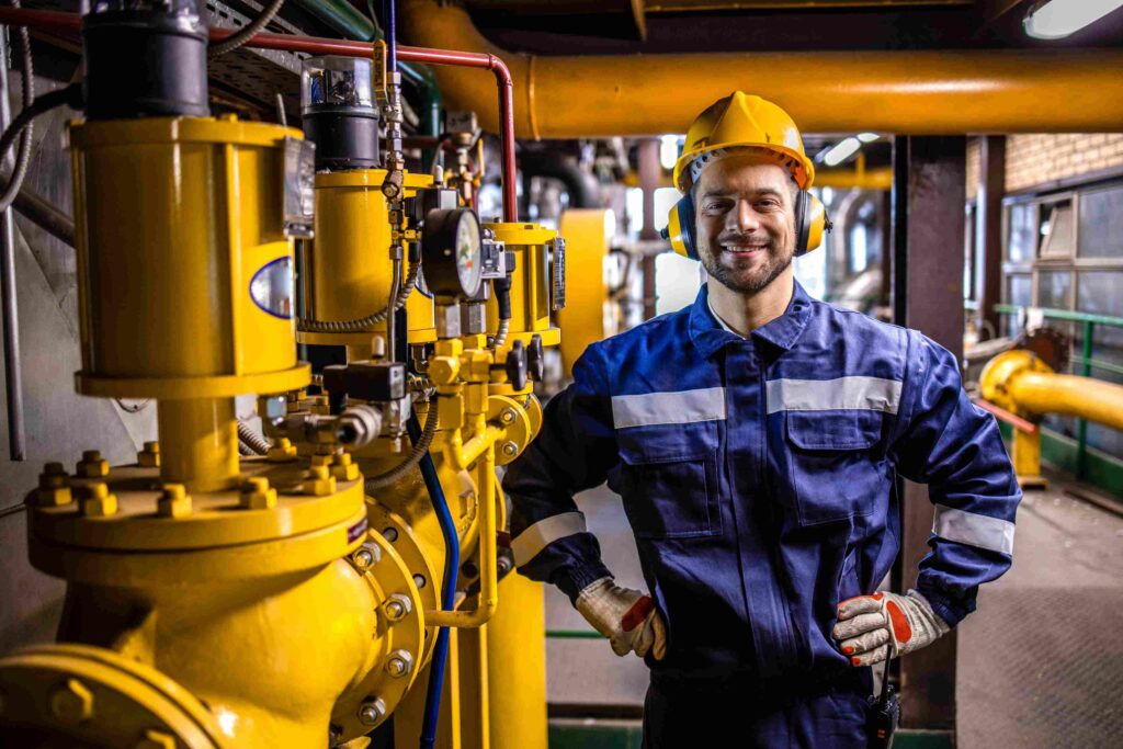 Ein Mechatroniker aus dem Ausland in blauer Schutzkleidung mit gelbem Helm arbeitet in einer Industrieanlage mit komplexen Maschinen.